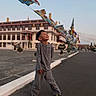 boy, building, casual, child, colorful, daylight, flag_banners, flags, grey_clothing, happy, joyful, outdoor, pavement, sky, smiling, standing, street, traditional_architecture, urban, yellow_shoes
