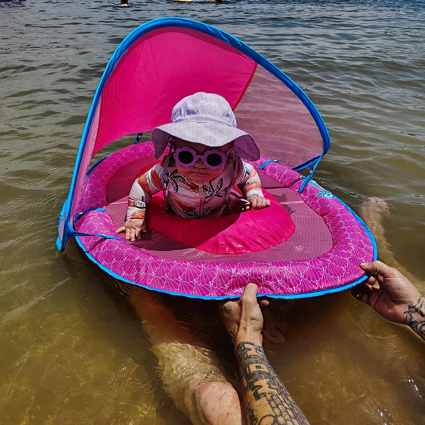Charli is registered to the contest to win money with this photo: adult, baby, canopy, child, float, floating_device, fun, hat, lake, nature, outdoor, person, pink, recreation, summer, sun_protection, sunglasses, tattoo, water, water_sport