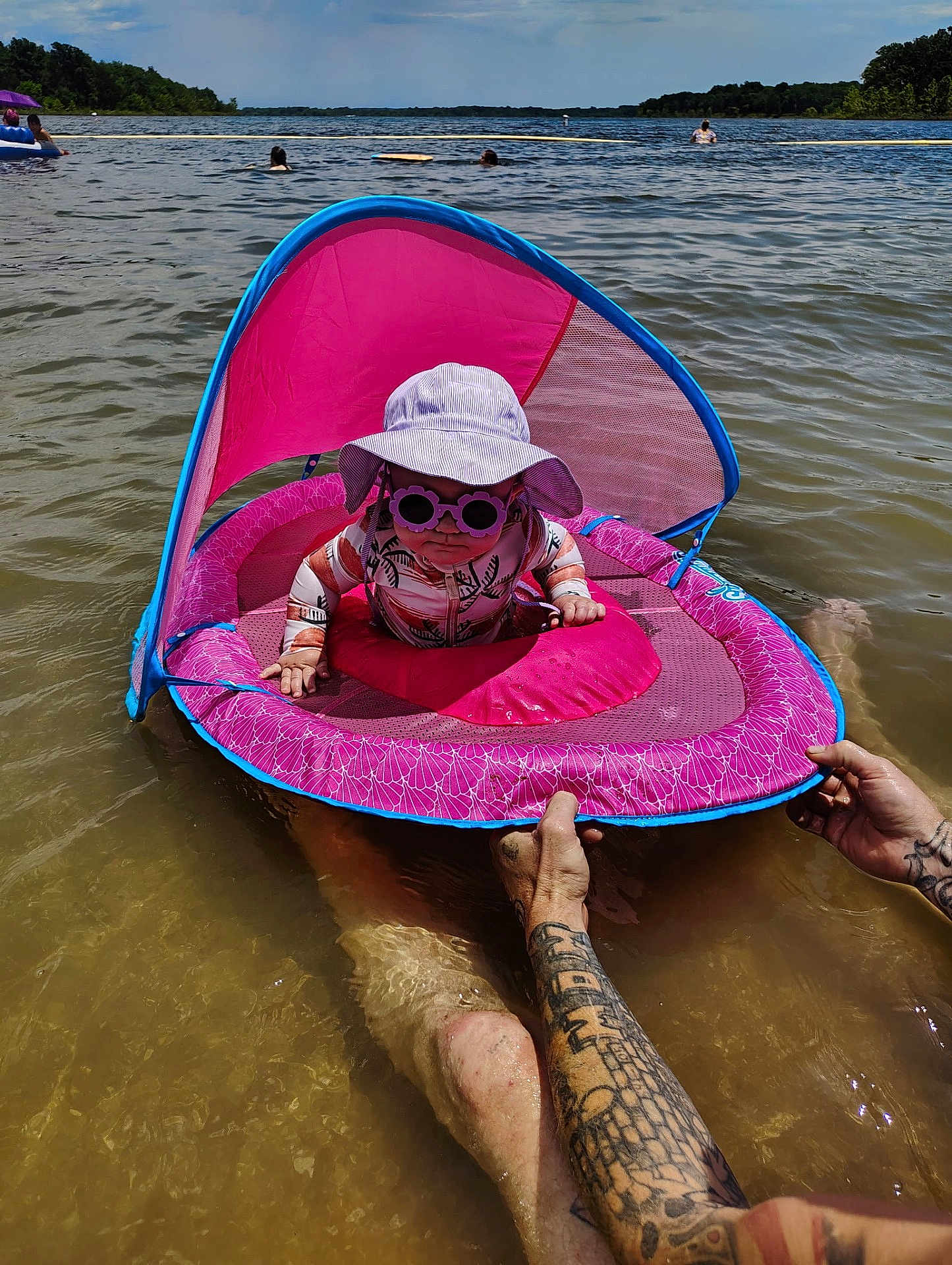 Charli is registered to the contest to win money with this photo: baby, child, float, water, lake, sunglasses, hat, sun_protection, tattoo, adult, summer, outdoor, recreation, fun, pink, canopy, floating_device, person, nature, water_sport