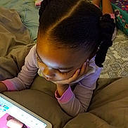 Anaya participe au concours pour gagner de l'argent avec cette photo : child, tablet, bed, blanket, braided_hair, stuffed_animal, indoor, sleepwear, person, technology, relaxing, cozy, casual, home, furniture, pink, screen, device, curled_hair, lying_down
