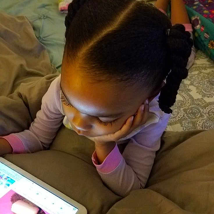 Anaya participe au concours pour gagner de l'argent avec cette photo : bed, blanket, braided_hair, casual, child, cozy, curled_hair, device, furniture, home, indoor, lying_down, person, pink, relaxing, screen, sleepwear, stuffed_animal, tablet, technology