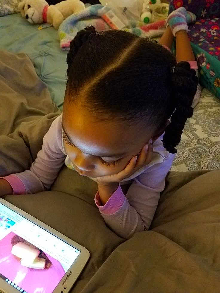 Anaya participe au concours pour gagner de l'argent avec cette photo : child, tablet, bed, blanket, braided_hair, stuffed_animal, indoor, sleepwear, person, technology, relaxing, cozy, casual, home, furniture, pink, screen, device, curled_hair, lying_down