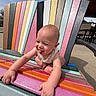 baby, child, smiling, tongue_out, colorful_chair, outdoor, sunlight, wood, pastel_colors, playful, happy, person, furniture, seat, cute, infant, summer, expression, fun, nature