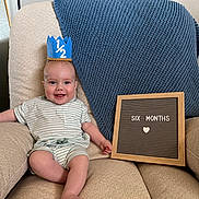 Charles is registered to the contest to win money with this photo: baby, infant, crown, chair, blanket, letter_board, smile, happy, milestone, sitting, clothing, striped_shirt, foot, hand, indoor, portrait, cute, child, celebration, home