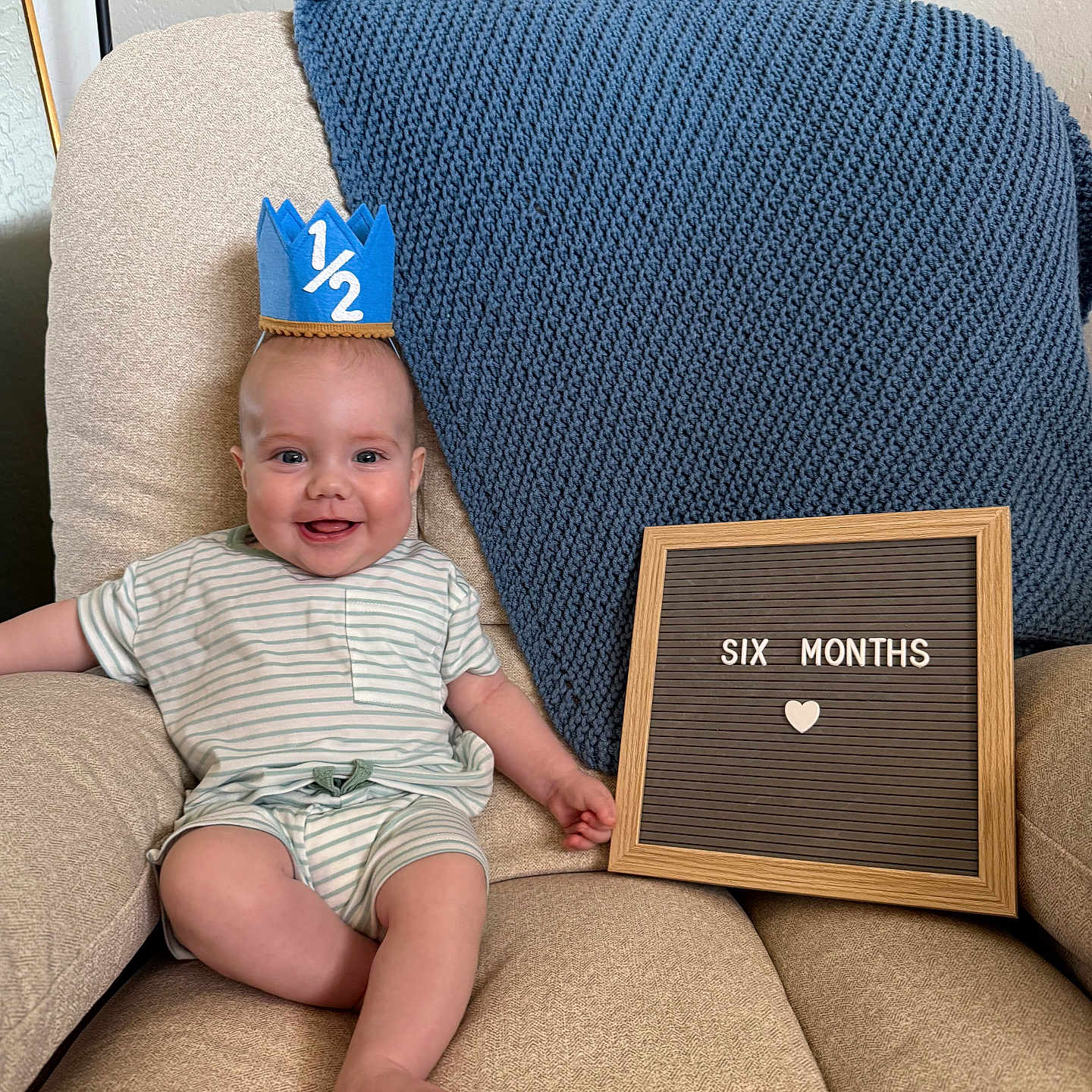 Charles is registered to the contest to win money with this photo: baby, infant, crown, chair, blanket, letter_board, smile, happy, milestone, sitting, clothing, striped_shirt, foot, hand, indoor, portrait, cute, child, celebration, home