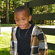 Jamerion is registered to the contest to win money with this photo: child, smiling, outdoor, plaid_jacket, black_shirt, fence, grass, sunlight, trees, happy, portrait, casual_clothing, earring, necklace, nature, young_child, person, daylight, standing, cute