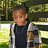 black_shirt, casual_clothing, child, cute, daylight, earring, fence, grass, happy, nature, necklace, outdoor, person, plaid_jacket, portrait, smiling, standing, sunlight, trees, young_child