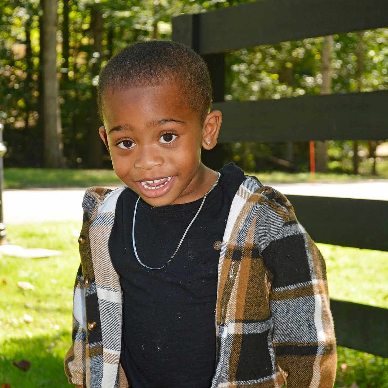 Jamerion is registered to the contest to win money with this photo: child, smiling, outdoor, plaid_jacket, black_shirt, fence, grass, sunlight, trees, happy, portrait, casual_clothing, earring, necklace, nature, young_child, person, daylight, standing, cute
