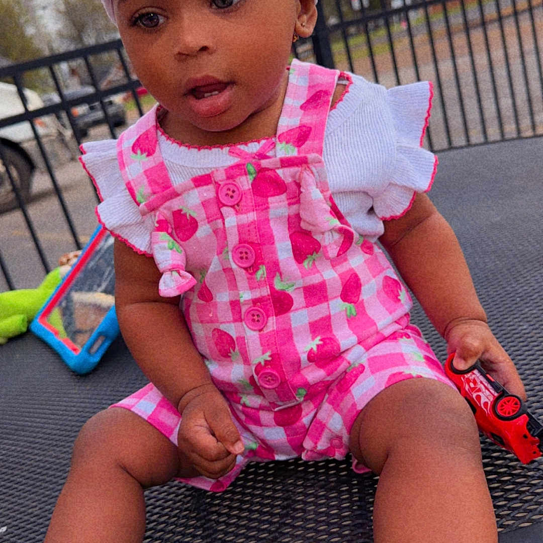 Harmony is registered to the contest to win money with this photo: toddler, child, headband, bow, romper, pink_clothing, toy_car, outdoor, table, metal_table, sitting, curious, cute, baby_shoes, footwear, person, daytime, play, expression, casual