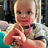 Calum joined the competition — help win amazing prizes! baby, child, infant, face, hands, string, chewing, clothing, indoors, furniture, sitting, expression, cute, room, shelf, blurred_background, skin, eye, playful, portrait