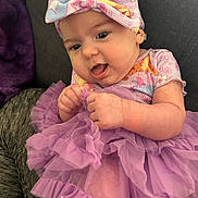 Annastasie is registered to the contest to win money with this photo: baby, infant, headband, bow, purple_tutu, dress, tulle, hands, tongue_out, cheeks, couch, plush_blanket, patterned_onesie, portrait, close_up, cute, smiling, fabric_texture, home_interior, sitting