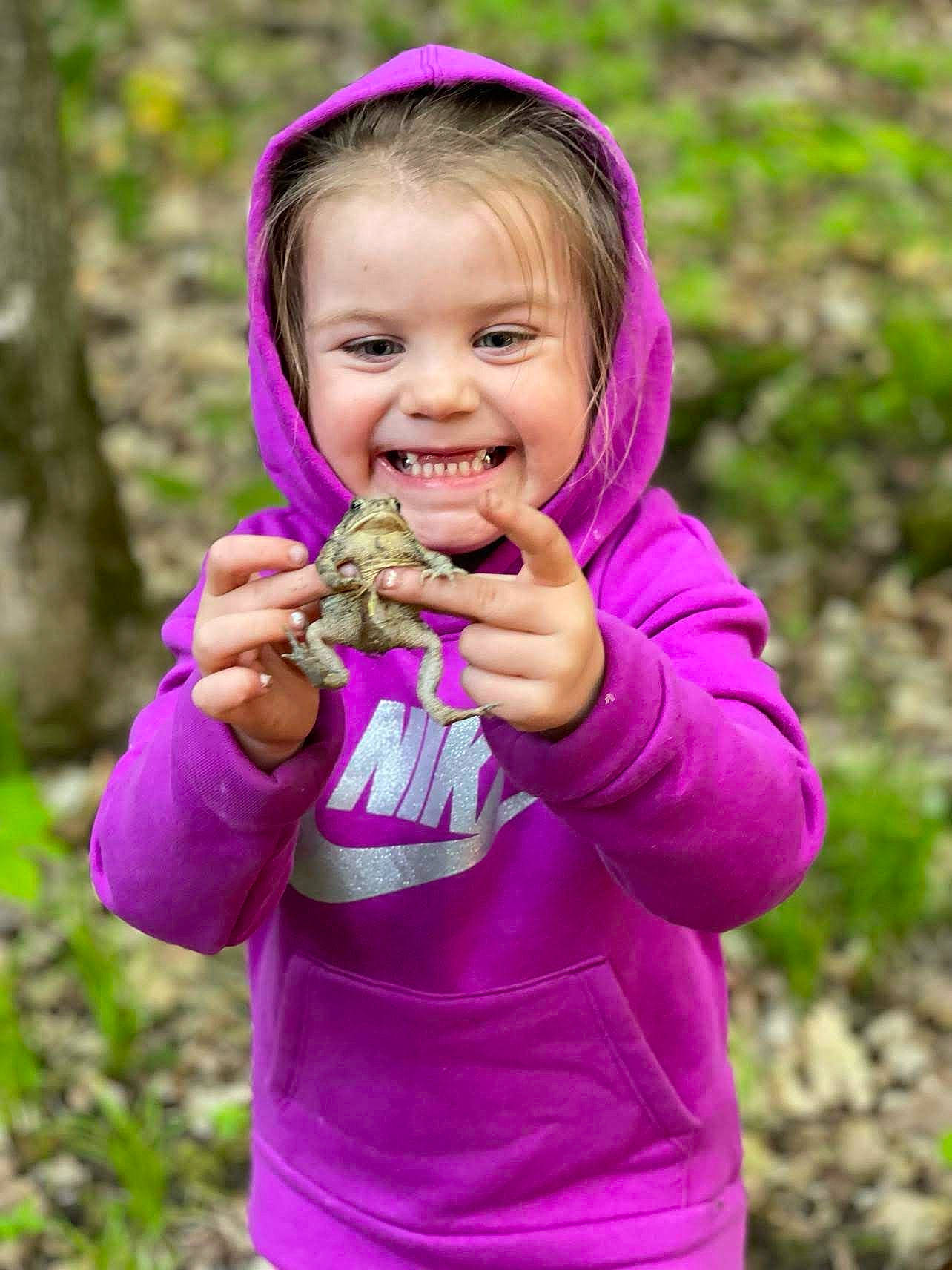 Gloria is registered to the contest to win money with this photo: arm, child, clothing, face, finger, fun, gesture, grass, hand, happy, joy, magenta, people_in_nature, person, pink, plant, purple, sleeve, smile, soil