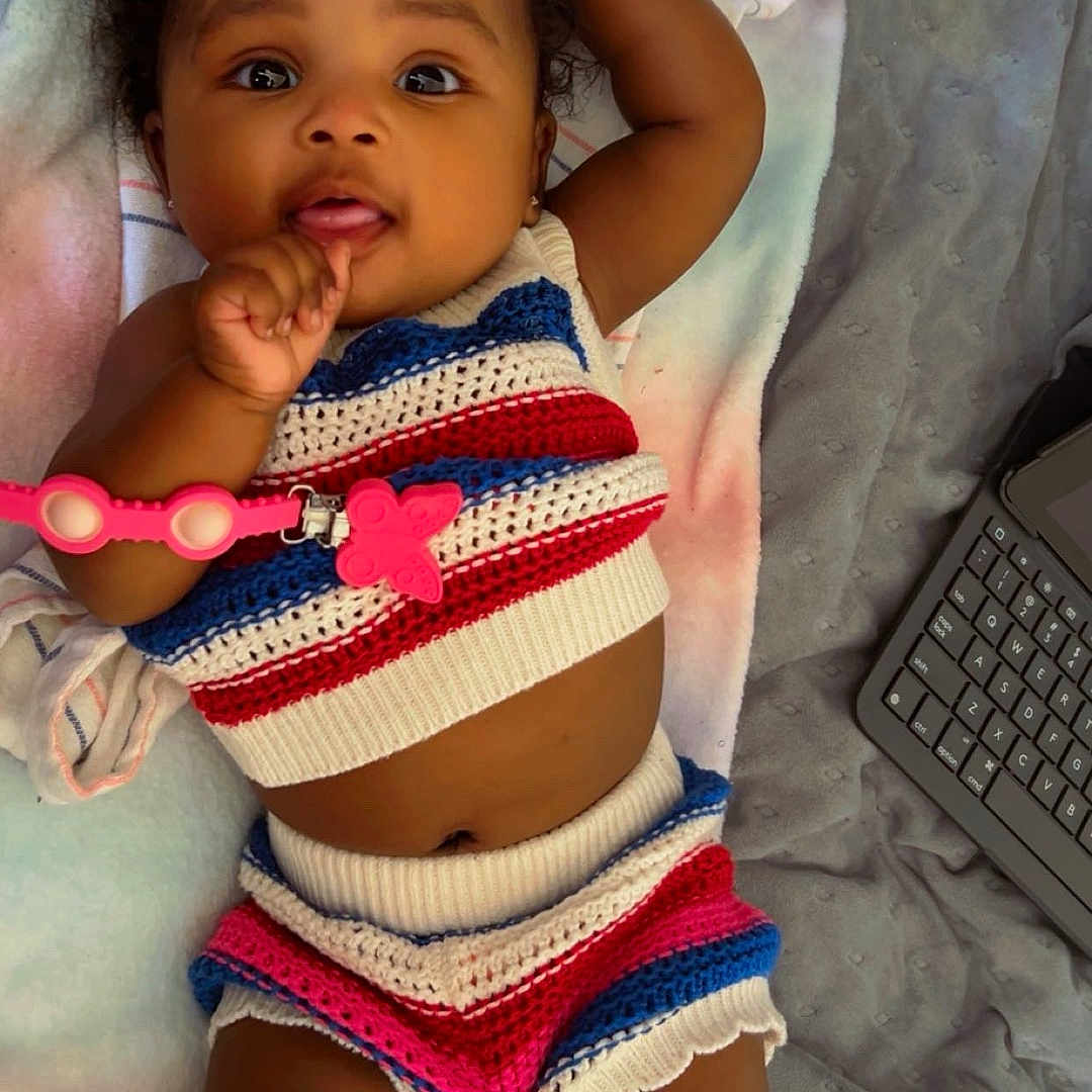 Sonyé is registered to the contest to win money with this photo: adorable, baby, blanket, bright_eyes, child, colorful_clothing, curly_hair, cute, hand_near_mouth, indoors, infant, keyboard, knitted_clothing, lying_down, pink_bow, portrait, relaxed, soft_surface, striped_outfit, young_child