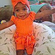 Kimora is registered to the contest to win money with this photo: baby, child, toddler, orange_clothing, headband, smiling, happy, arms_outstretched, cushion, floral_pattern, indoor, living_room, sofa, person, cute, seated, home, bright_colors, portrait, infant