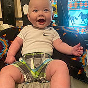 Josiah joined the competition — help win amazing prizes! baby, smiling, infant, sitting, white_shirt, plaid_shorts, colorful_cushion, indoor, happy, cute, child, patterned_fabric, wooden_beams, curtains, bright, cheerful, portrait, young_child, clothing, room