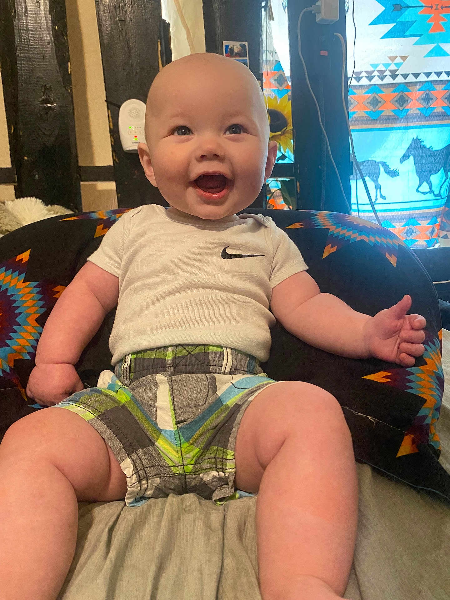 Josiah joined the competition — help win amazing prizes! baby, smiling, infant, sitting, white_shirt, plaid_shorts, colorful_cushion, indoor, happy, cute, child, patterned_fabric, wooden_beams, curtains, bright, cheerful, portrait, young_child, clothing, room