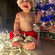 Josiah is registered to the contest to win money with this photo: baby, child, christmas, holiday, lights, santa_hat, red_pants, smiling, indoors, floor, decorated_tree, gift_box, festive, cute, toes, holiday_decor, joyful, celebration, toddler, christmas_tree