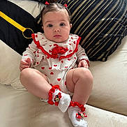 Italy is registered to the contest to win money with this photo: baby, infant, child, couch, pillow, heart_pattern, white_onesie, red_bows, socks, minnie_mouse, cute, curious, indoor, portrait, person, sitting, hair_ties, soft_fabric, decor, plush