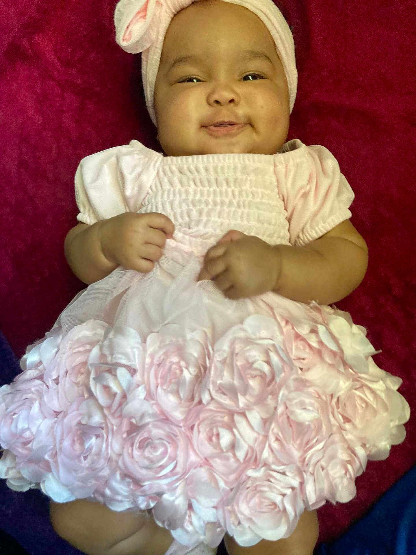 Lailani Anaisa is registered to the contest to win money with this photo: baby, child, pink_dress, headband, smile, roses, frilly, cute, infant, cheeks, fabric, clothing, portrait, lying_down, adorable, happy, toddler, hands, background, red