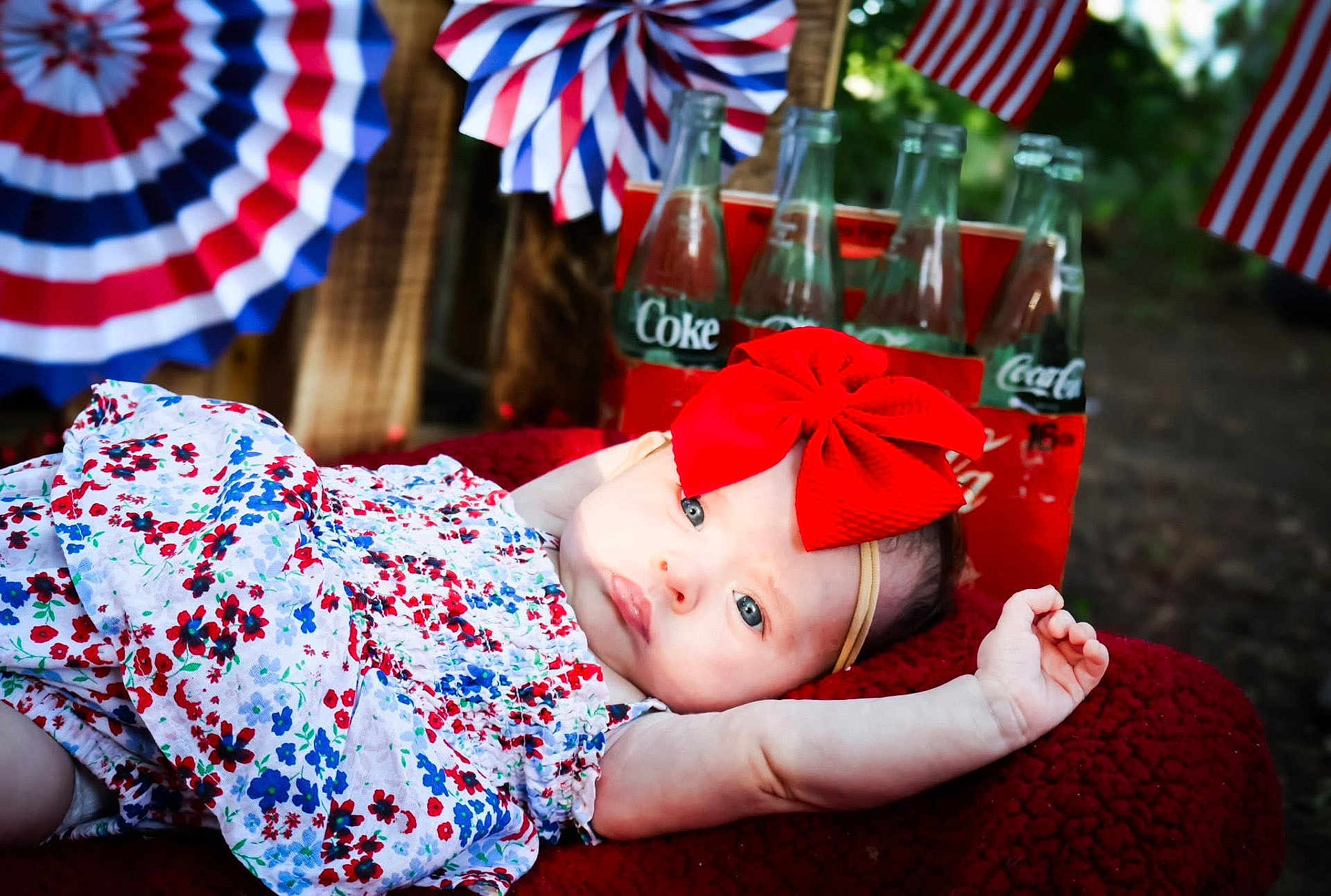 Avery is registered to the contest to win money with this photo: baby, infant, red_bow, blue_eyes, floral_dress, blanket, coca_cola, glass_bottles, patriotic, flags, decorations, outdoor, portrait, cute, celebration, summer, relaxing, holiday, headband, person