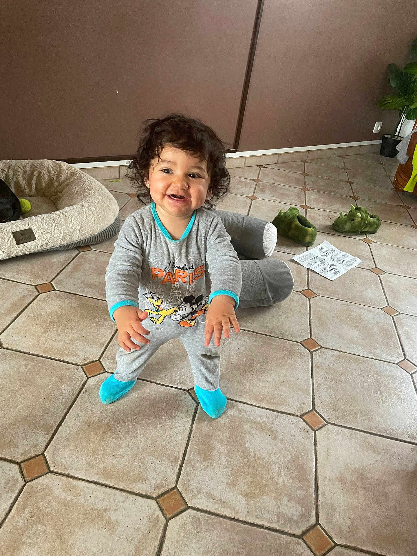 Reyna participe au concours pour gagner de l'argent avec cette photo : toddler, child, smiling, curly_hair, onesie, disney, mickey_mouse, pluto, blue_socks, indoor, floor_tiles, dog_bed, slippers, dinosaur_slippers, paper, plant, brown_wall, happy, standing, home