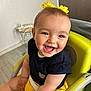 baby, child, smiling, happy, high_chair, yellow, hair_accessory, flower, clothing, black_shirt, yellow_shorts, indoor, person, cute, face, skin, arm, teeth, seat, joy