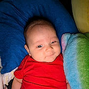 Liam joined the competition — help win amazing prizes! baby, infant, red_clothing, pillow, blanket, soft_texture, smile, face, head, comfort, resting, cute, child, indoor, cozy, colorful, lying_down, young, person, portrait