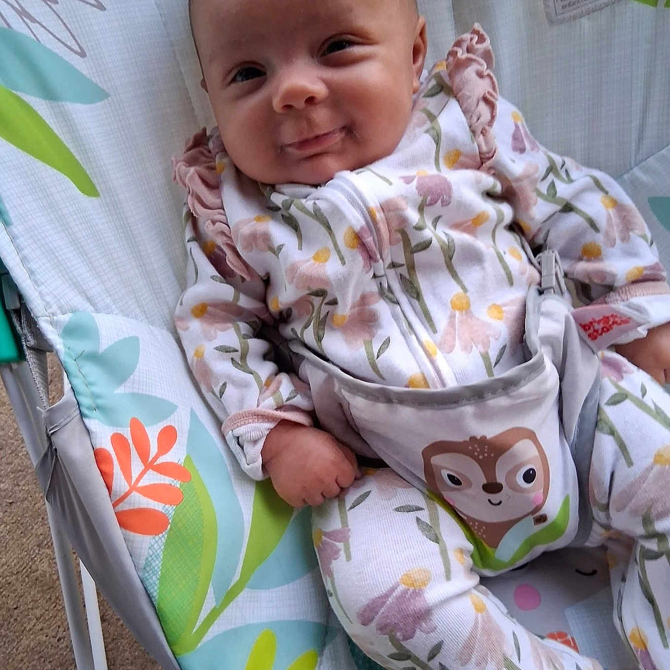 Raya is registered to the contest to win money with this photo: baby, baby_seat, child, clothing, colorful, comfort, cute, expression, face, floral_pattern, hand, indoor, infant, legs, onesie, pattern, playful, resting, seat, smiling