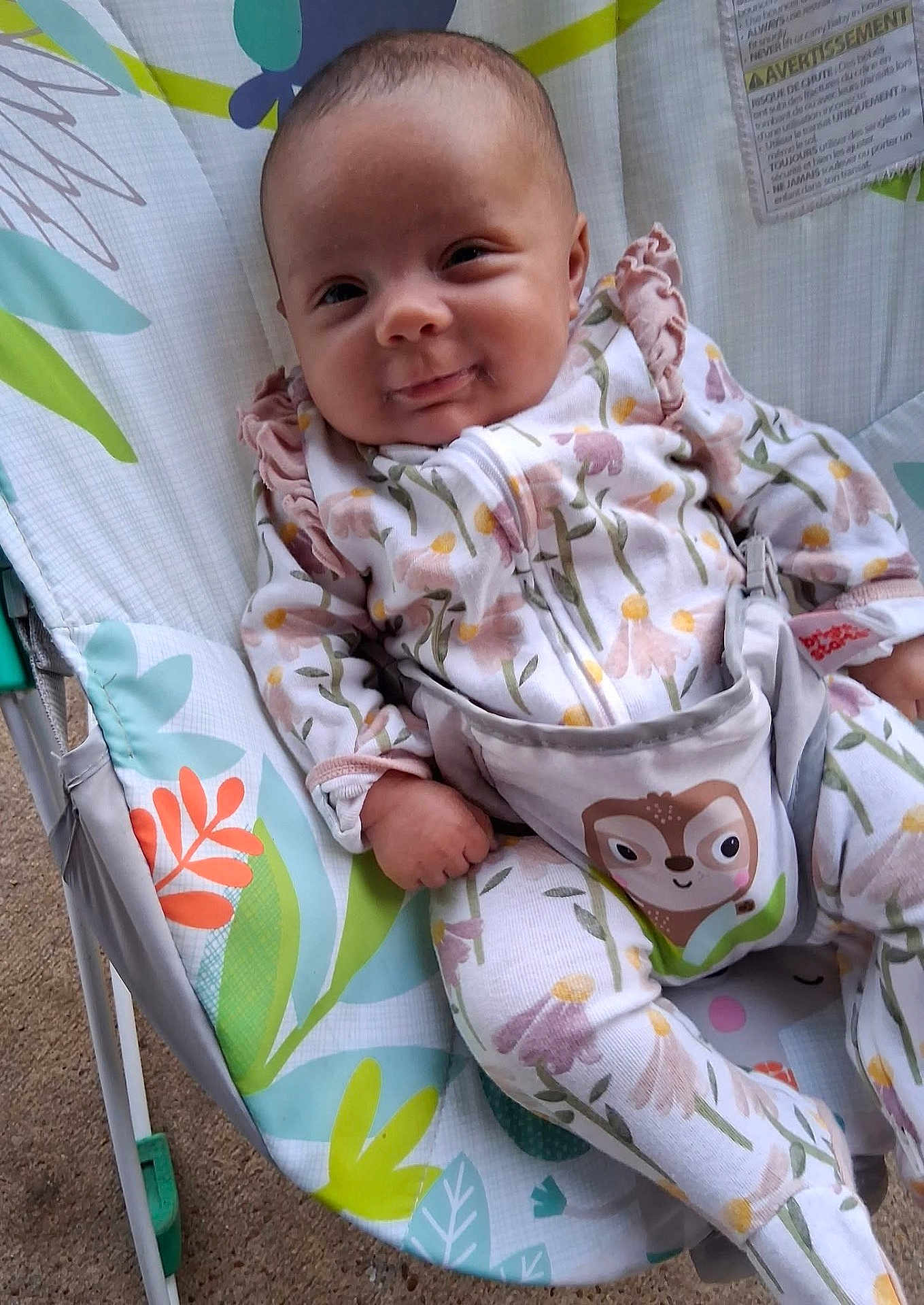 Raya is registered to the contest to win money with this photo: baby, infant, onesie, floral_pattern, baby_seat, smiling, cute, child, face, hand, legs, comfort, indoor, clothing, seat, playful, expression, pattern, colorful, resting