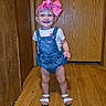 toddler, child, baby, smiling, standing, portrait, pink_bow, hair_accessory, denim_overalls, shirt, sandals, indoor, hallway, wood_paneling, wooden_floor, door, home, happy, full_body, photograph