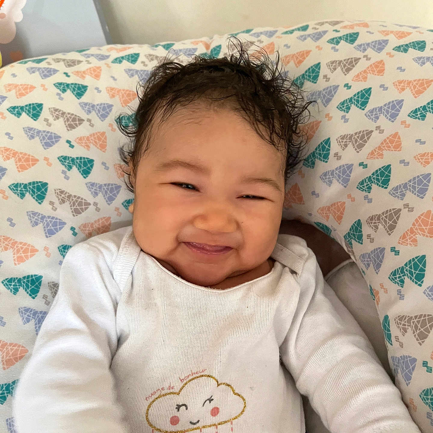 Monie participe au concours pour gagner de l'argent avec cette photo : baby, cheeky_expression, child, closeup, cloud_design, colorful_pattern, cozy, curly_hair, cute, happy, heart_pattern, indoors, infant, lying_down, person, pillow, smiling, soft_lighting, white_onesie, young_child
