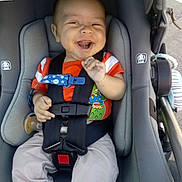 Angel is registered to the contest to win money with this photo: baby, car_seat, smiling, child, orange_shirt, dinosaur_print, seatbelt, happy, infant, cute, hands, clothing, safety_harness, casual, outdoor, daylight, footwear, pants, person, expression