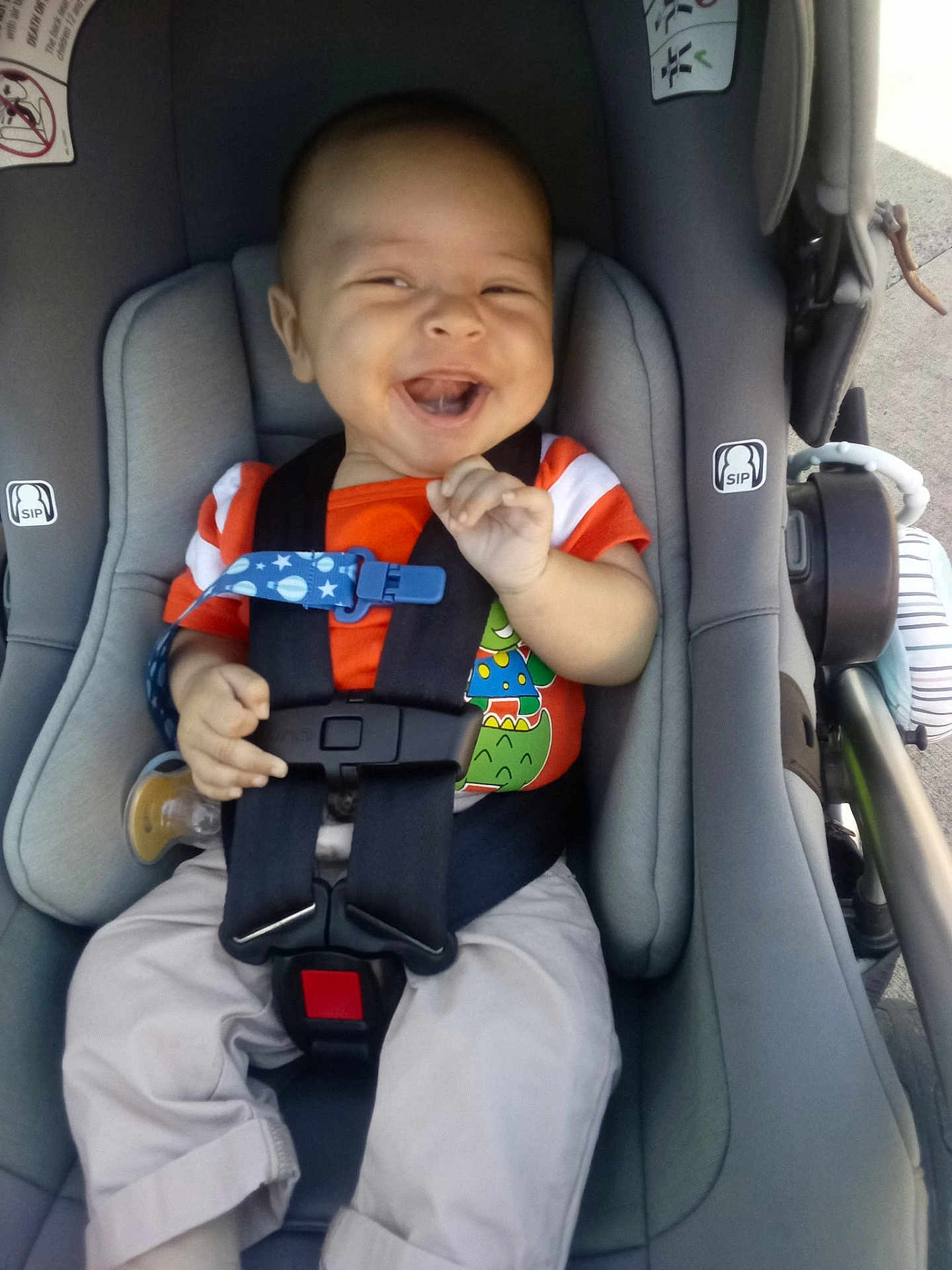 Angel is registered to the contest to win money with this photo: baby, car_seat, smiling, child, orange_shirt, dinosaur_print, seatbelt, happy, infant, cute, hands, clothing, safety_harness, casual, outdoor, daylight, footwear, pants, person, expression