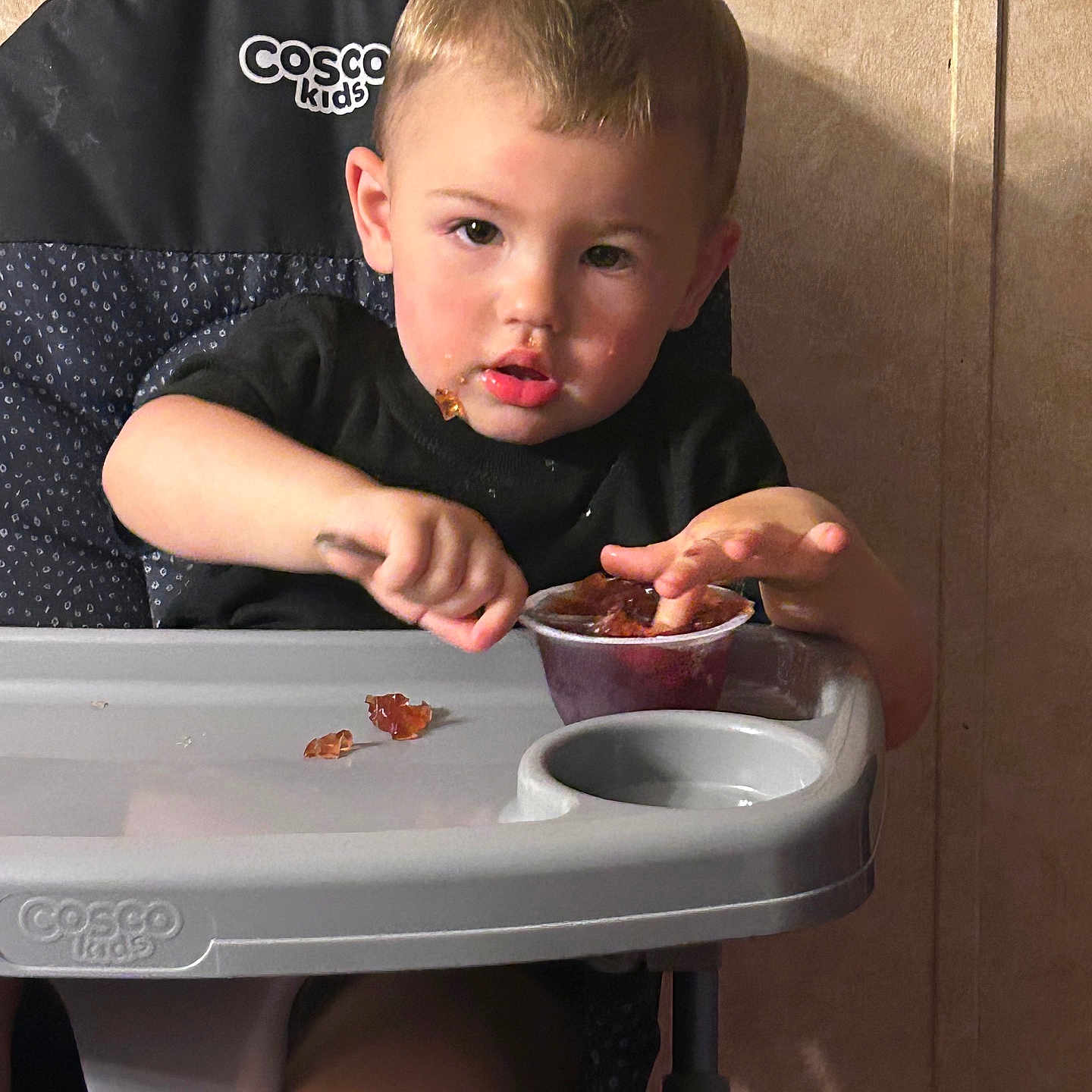 Zayden is registered to the contest to win money with this photo: black_shirt, child, concentration, cosco_kids, cup, eating, expression, face, food, high_chair, indoor, jelly, mealtime, mess, messy, person, plastic_tray, spoon, toddler, young_child