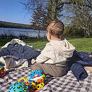 Paolo participe au concours pour gagner de l'argent avec cette photo : child, baby, back_view, head, hair, blanket, picnic, toys, toy_ball, rattle, playmat, lake, water, trees, grass, outdoor, sunny, jacket, hoodie, plaid_blanket