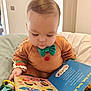 baby, infant, child, board_book, reading, onesie, bow_tie, hand, toy, pillow, bedding, indoors, closeup, face, hair, eyelashes, colorful, cute, sitting, cozy