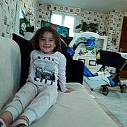 Ambre participe au concours pour gagner de l'argent avec cette photo : child, smiling, couch, living_room, curly_hair, socks, pajamas, toy, stroller, television, wallpaper, pillow, plant, lamp, footwear, furniture, indoor, portrait, playful, home