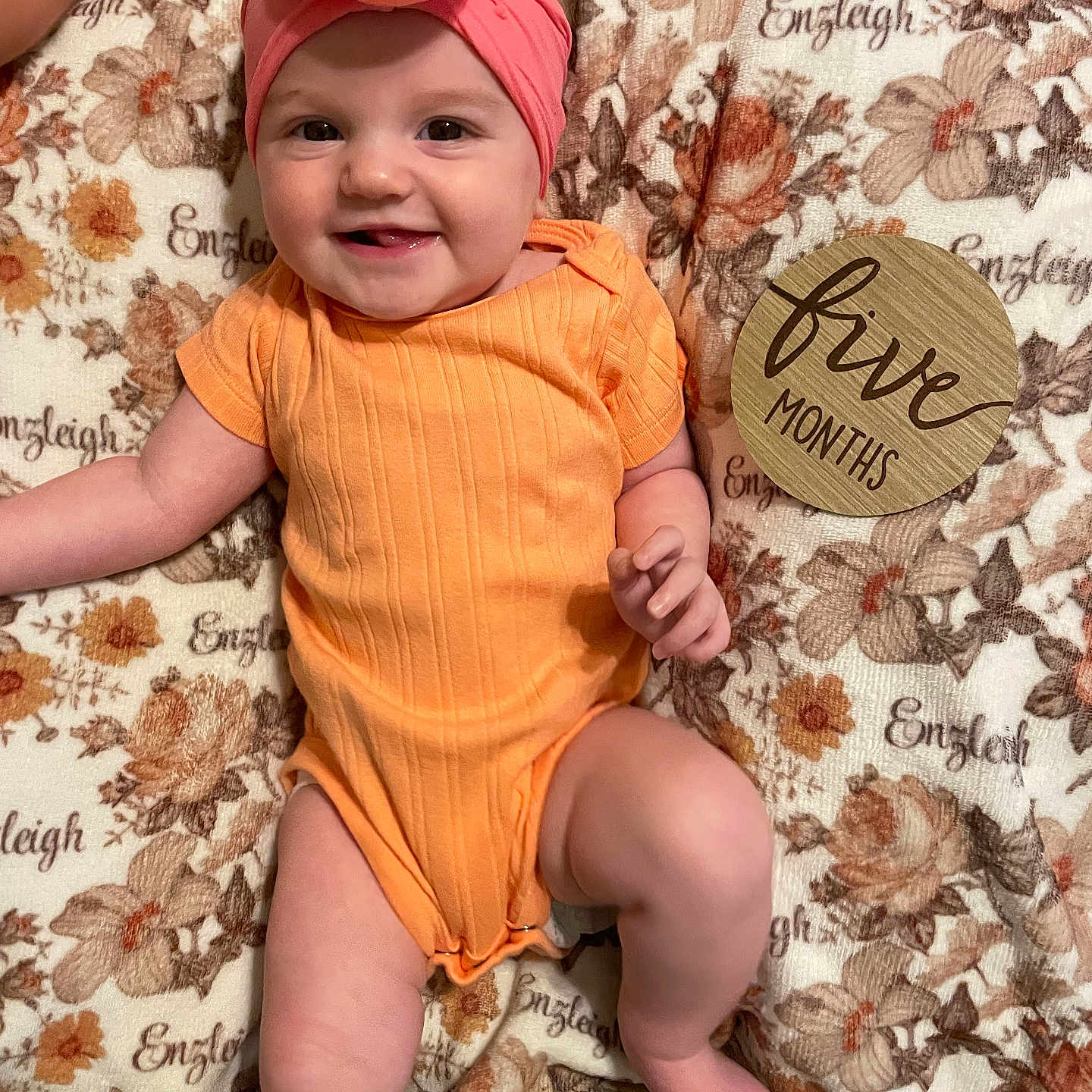 Enzleigh is registered to the contest to win money with this photo: baby, child, chubby_legs, cute, donut_shape, five_months, floral_blanket, hand, happy, headband, human, indoors, infant, lying_down, orange_onesie, pink_headband, portrait, skin, smiling, wooden_sign