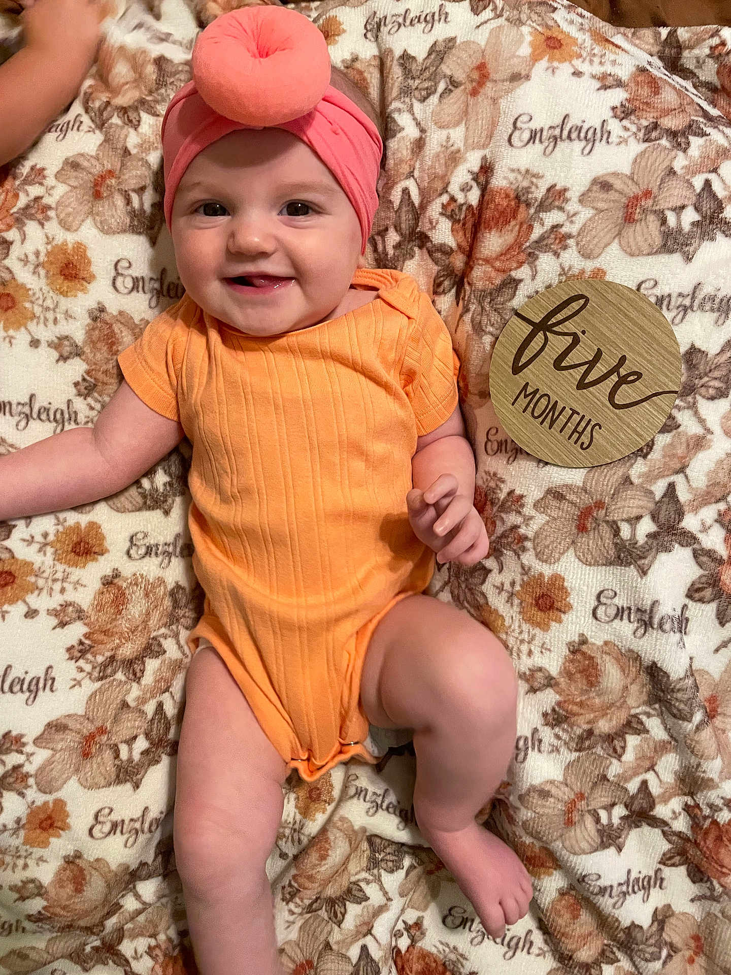 Enzleigh is registered to the contest to win money with this photo: baby, smiling, infant, orange_onesie, headband, pink_headband, donut_shape, floral_blanket, wooden_sign, five_months, cute, chubby_legs, happy, lying_down, indoors, child, portrait, human, skin, hand