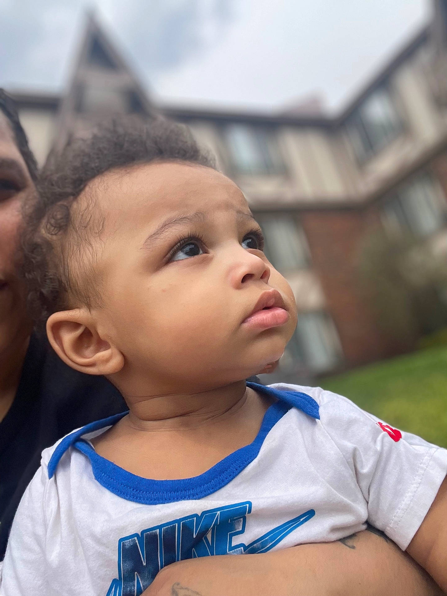 Silas is registered to the contest to win money with this photo: adult_hand, background_blur, blue_eyes, building, casual_clothing, child, closeup, curly_hair, daylight, expression, face, grass, nike_logo, outdoor, person, portrait, skin, toddler, white_shirt, young_child