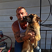 Dobby is registered to the contest to win money with this photo: man, dog, tattoos, beard, porch, sunlight, shadow, casual, pet, companion, jeans, tank_top, metal_furniture, ashtray, cigarette_butts, can, wood_floor, wall, outdoor, friendship