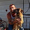 ashtray, beard, can, casual, cigarette_butts, companion, dog, friendship, jeans, man, metal_furniture, outdoor, pet, porch, shadow, sunlight, tank_top, tattoos, wall, wood_floor