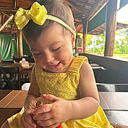 Julie a rejoint le concours — aidez-le/la à gagner de superbes lots ! baby, bottle, bow, casual, chair, child, curious, cute, happy, headband, holding, indoor, infant, natural_light, person, portrait, smiling, table, wooden_structure, yellow_dress