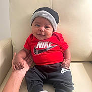 Deangelo is registered to the contest to win money with this photo: baby, child, smile, nike, hat, red_shirt, black_pants, socks, hand, chair, leather, indoor, portrait, cute, person, infant, clothing, comfort, seated, happy