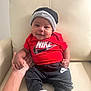 baby, child, smile, nike, hat, red_shirt, black_pants, socks, hand, chair, leather, indoor, portrait, cute, person, infant, clothing, comfort, seated, happy