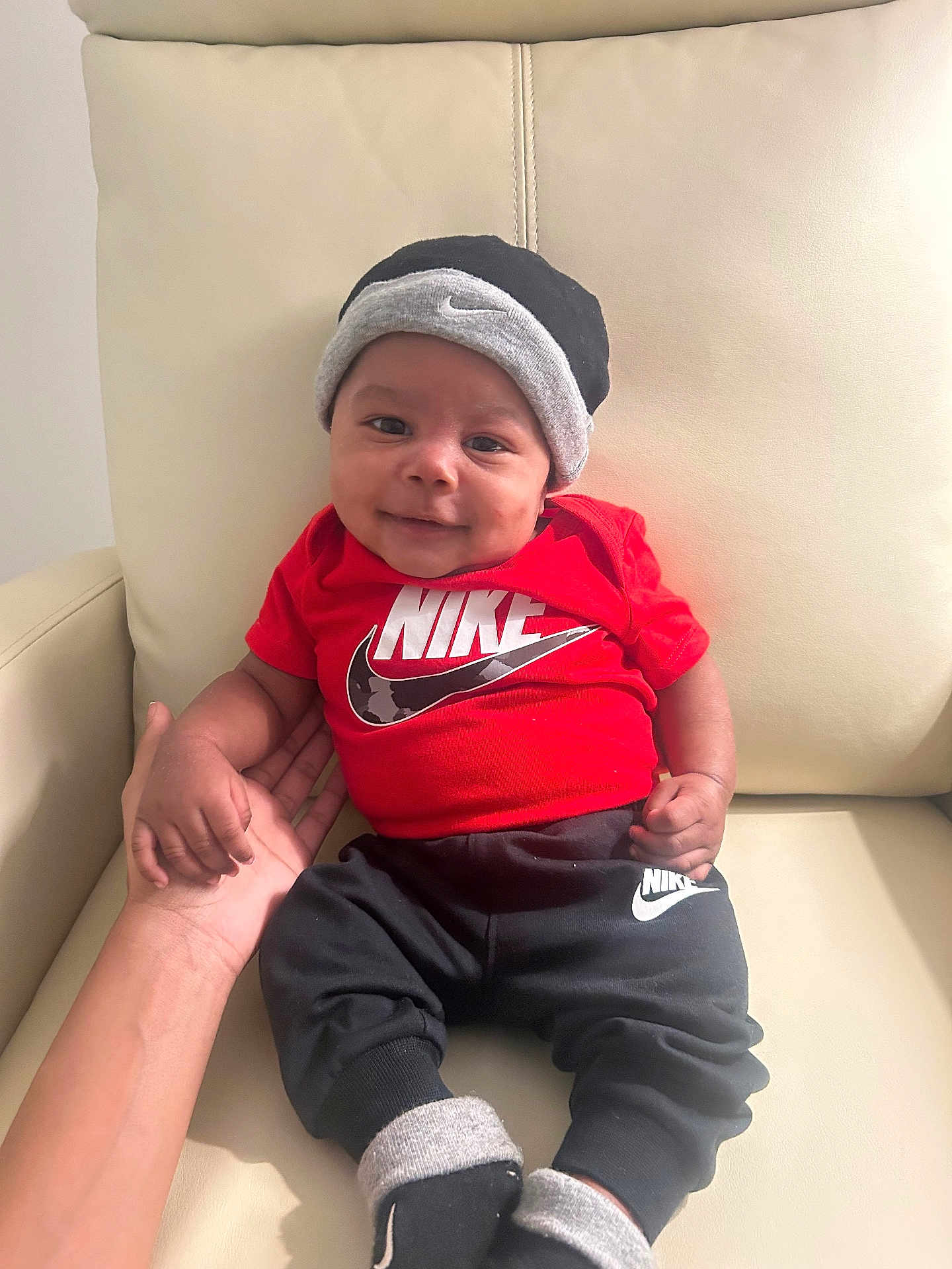 Deangelo is registered to the contest to win money with this photo: baby, child, smile, nike, hat, red_shirt, black_pants, socks, hand, chair, leather, indoor, portrait, cute, person, infant, clothing, comfort, seated, happy