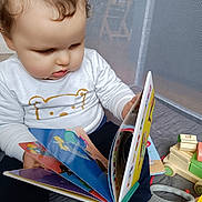 Maturin participe au concours pour gagner de l'argent avec cette photo : baby, board_book, book, child, colorful, curly_hair, early_learning, face, floor_mat, focused, hand, indoor, playpen, playtime, reading, sitting, toddler, toy, white_shirt, wooden_toy
