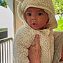baby, child, infant, onesie, hood, ears, fluffy_clothing, hand, freckles, indoor, window, natural_light, curious_expression, surprised_face, person, portrait, cute, soft_texture, warm_clothing, holding