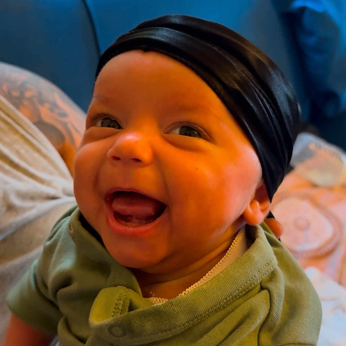 J Pierce joined the competition — help win amazing prizes! adult_hand, baby, blue_couch, child, clothing, couch, cute, expression, green_onesie, happy, headwrap, home, indoor, infant, laughing, person, portrait, smiling, tattoo, window