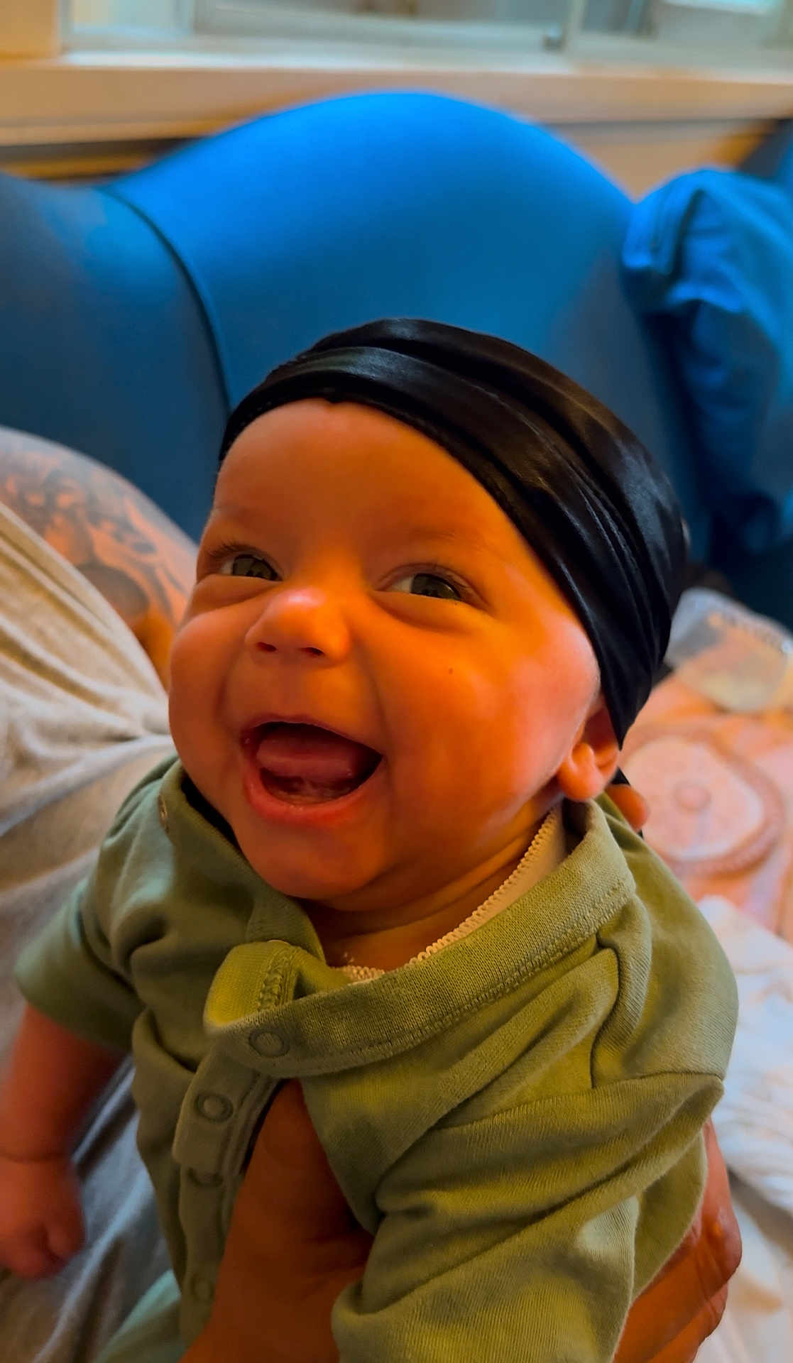 J Pierce joined the competition — help win amazing prizes! baby, child, smiling, laughing, headwrap, green_onesie, tattoo, adult_hand, indoor, couch, blue_couch, window, person, happy, cute, portrait, infant, clothing, expression, home